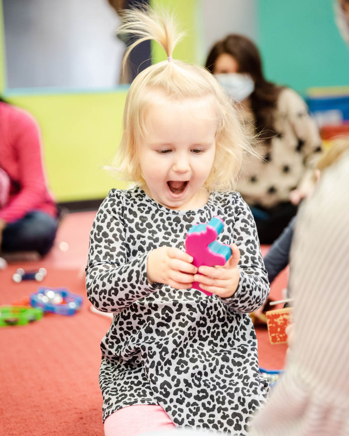 A littel girl with blonde hair holding a toy and smiling at Romp n' Roll in Willow Grove.