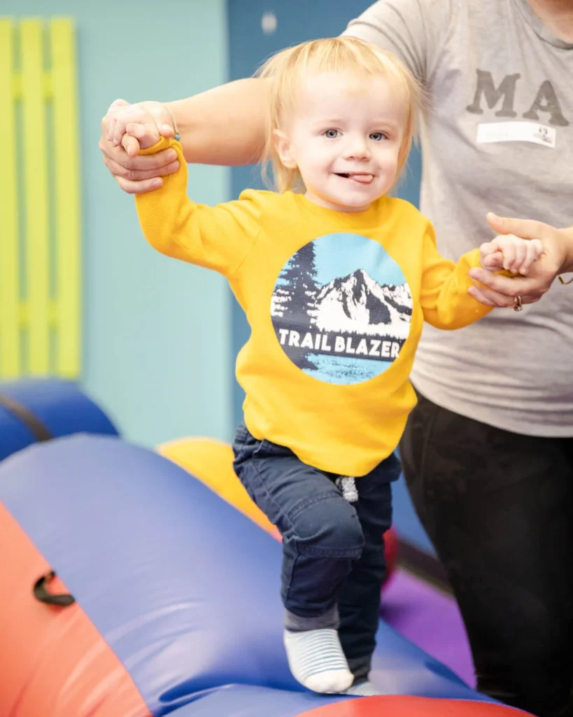 A young toddler playing on gym equipment with the help of a Romp n' Roll staff member.