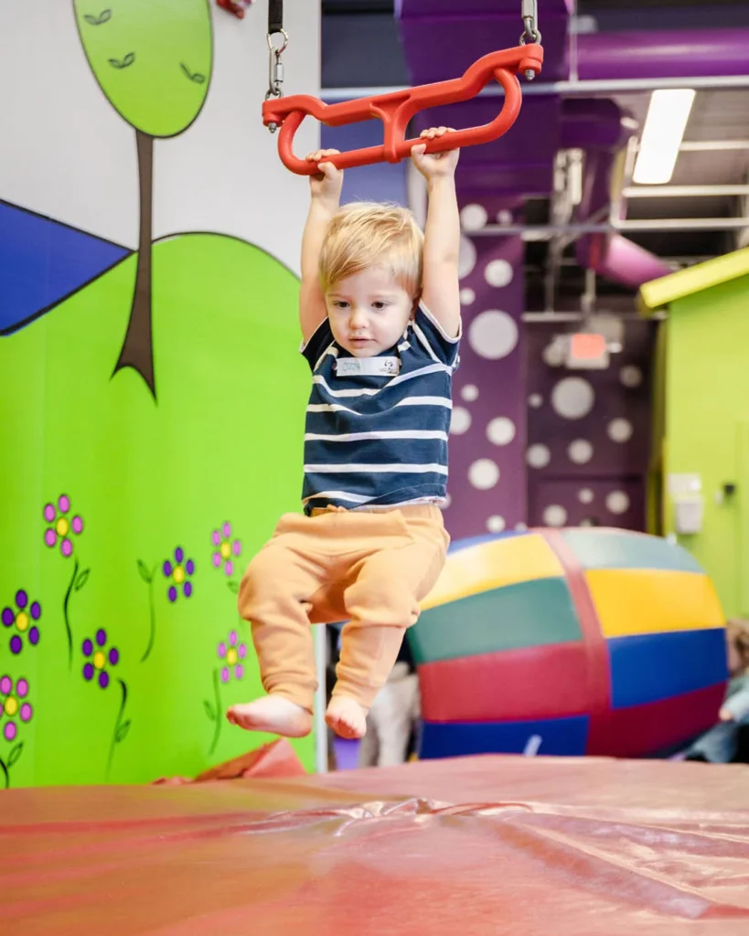 A young boy hanging from a bar while playing at a Romp n' Roll gym.