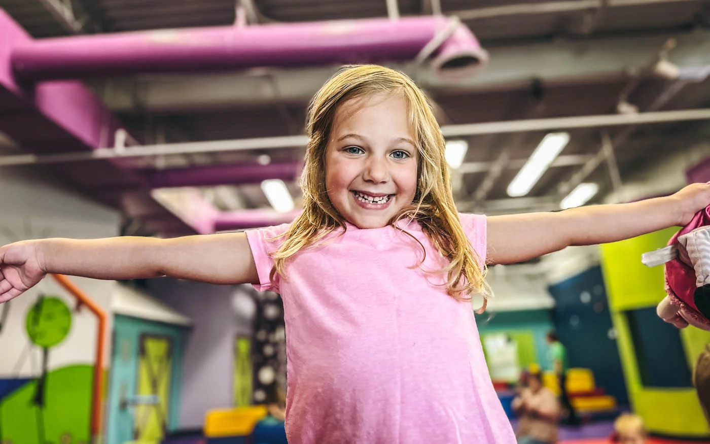 A young girl holds her arms out and smiles for the camera as she plays at Romp n' Roll, a kids gym franchise.